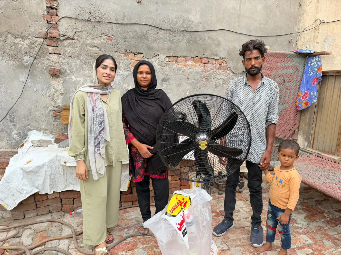 Three adults and a child standing beside a large electric fan in a courtyard