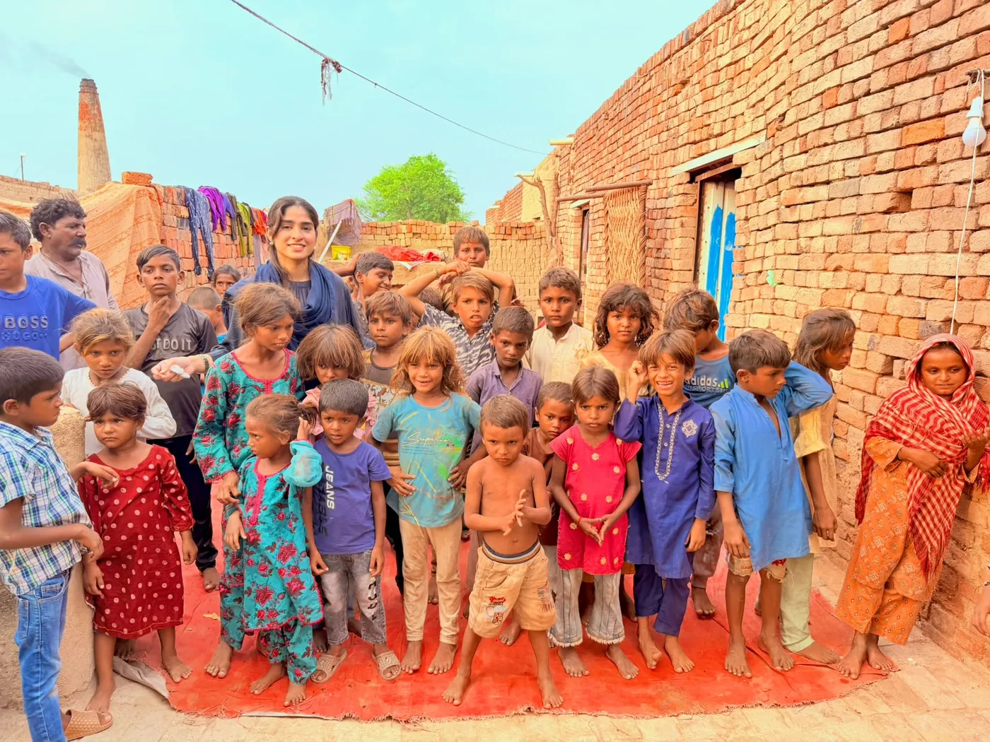 A group of children and adults gathered on a red mat beside brick buildings