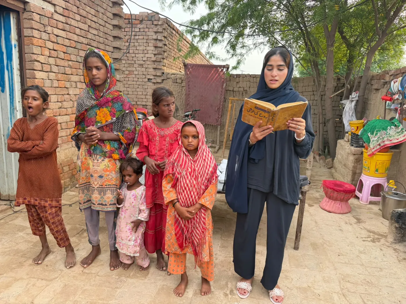 A woman reading from a book alongside children in a courtyard