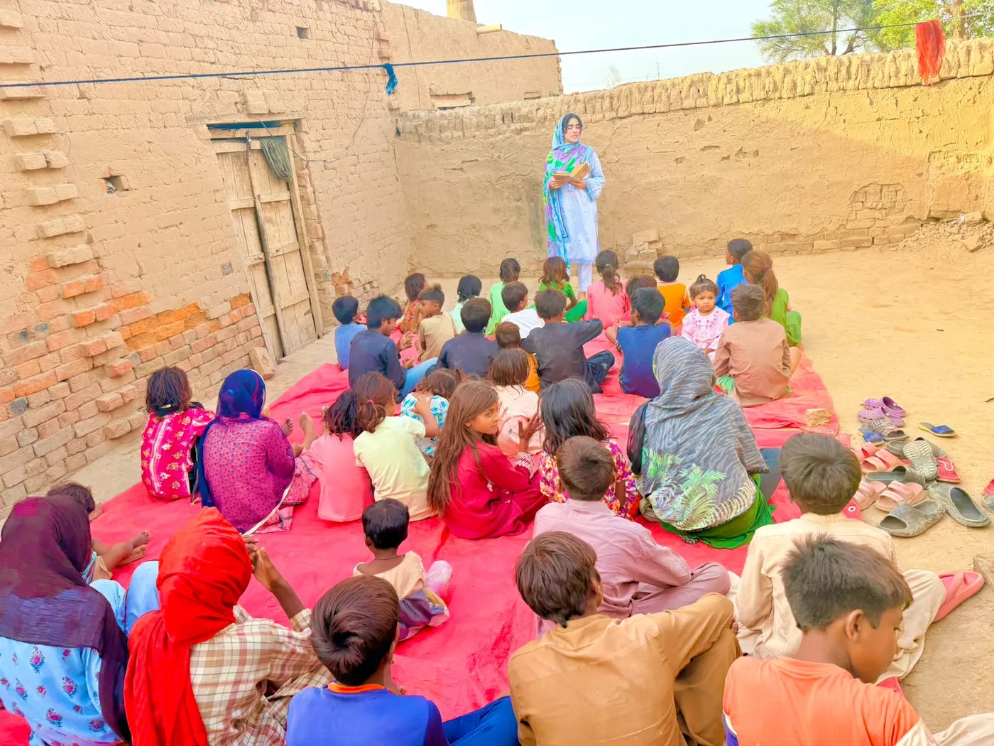 A group of children seated on a red mat facing a woman outdoors