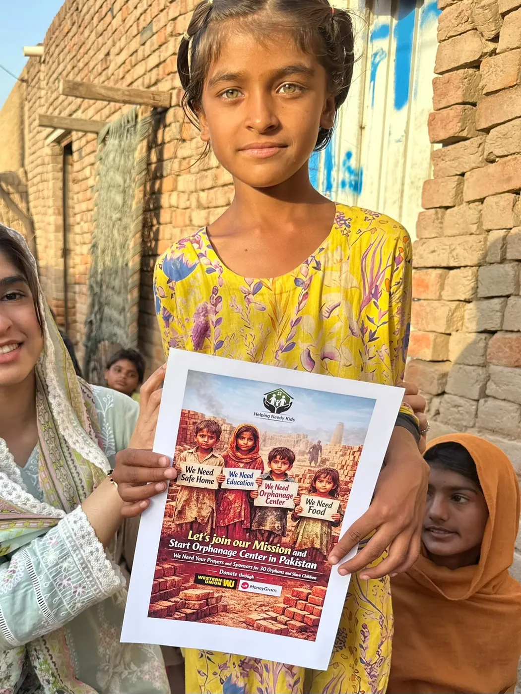 A young girl holding a printed flyer beside a brick wall with other people nearby